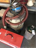 Shop vacuum with coiled red hose and electrical cord, blue upper motor cover with label, silver collection tank on wheels.