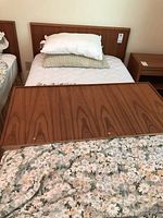 Full setup showing single bed with teak laminate headboard and metal rails, mattress, pillows, floral comforter, and box spring. Second smaller headboard displayed on bed.