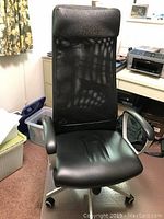 Front view of black office chair showing high mesh back, leather headrest, leather seat, armrests, and rolling base.