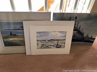 Three art pieces grouped on carpet, including framed landscape watercolour, unframed watercolour, and unframed photograph.