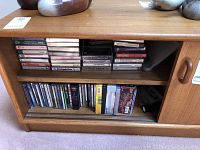 Wooden shelf with two rows of cassette tapes on the top shelf and DVDs/CDs on the bottom shelf. Various classical music titles and artists visible on cassette spine labels.