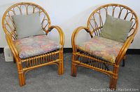 Frontal view of the matched pair of vintage rattan chairs with floral seat cushions and green throw pillows on a gray carpet.