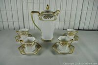 Full set photo showing one octagonal chocolate pot with lid and four hexagonal cups and saucers decorated with gold trim and floral pattern.