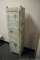 Front angled view of tall folk art painted cupboard showing hot air balloons on the door and sides, decorative round knobs on top and door.