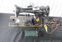 Full view of black vintage Singer sewing machine with rotary attachments box in front, showing power cord and foot pedal.