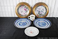 Photo showing all six items arranged on a black surface: two framed hummingbird plates in gold frames at the back, two blue and white Canton plates in front, and the Wedgwood Peter Rabbit mug and plate centered in front.