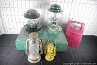 Photo showing two Coleman lanterns, one missing globe; two barn lanterns (silver and yellow); green metal carrying case and red Coleman plastic carrying case.