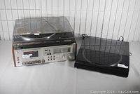Photo of the three stereo components on a white surface against a tiled white background: cassette deck with wooden side panels on bottom left, linear turntable with dust cover on right, and standard turntable on top of cassette deck.