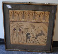 Framed textile needlework with a central scene of a nomad riding a donkey and pulling a camel, with palm trees and decorative border, showing complete piece within frame.