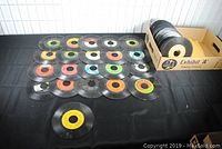 Top-down photo showing 24 vintage 45rpm records with varied colored center labels arranged on black surface and additional records in a cardboard box.
