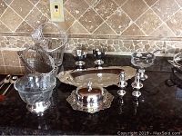 Overview of silverplate and chrome/stainless steel serving pieces on dark countertop showing bowls, baskets, trays, covered butter dish, salt and pepper shakers.