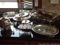 Variety of silver plated serving pieces arranged on counter, including chafing dish, glass tray, covered butter dish, spoons, candle snuffer.