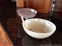 Two bowls on dark speckled granite countertop: a clear glass footed bowl with red border and a round alabaster design bowl.