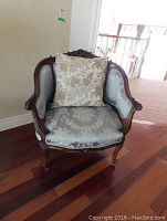 Front view of the carved wood armchair with blue damask fabric and floral pillow sitting on the seat