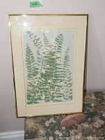 Framed art print featuring green fern leaves numbered 2/12 with illegible signature, standing on a floral patterned surface against plain wall.