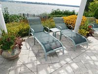 Two green mesh outdoor chaise lounges and small side table arranged on stone patio by water view