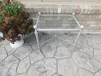 Front angle of outdoor glass and metal side table on stamped concrete patio next to brick wall and planter.