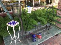 Photo showing white and blue ceramic tile top plant stand, tall iron and glass bulb planter, black wire shelf stand, and glass watering balls on a table with a fern background.