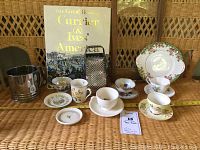 Wide view of tea lot with all items displayed on wicker surface: metal sifter, metal grater, Royal Stafford Olde English Garden cake plate, glass and wood tray, assorted teacups and saucers, and large Currier & Ives America coffee table book.