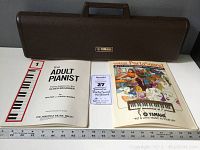Brown Yamaha hard carrying case placed on gray surface with three books and guides in front, measuring approx 20 inches in length. Books include 'The Adult Pianist' piano basics for older beginners, Yamaha PortaSound playing guide, and a sheet music page.