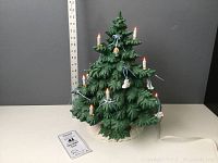 Full view of green ceramic Christmas tree decorated with candle lights and plaster ornaments on a white ceramic base.