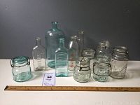 Lot of 11 vintage glass bottles and canning jars displayed on table with ruler for size reference, showing variety of shapes, colors and closures.