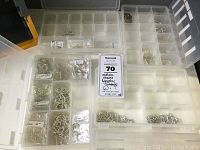 Top view of divided clear plastic storage case opened containing many small compartments filled with silver-colored metal jewelry supplies including jump rings, chains, and possibly clasp components and earring hooks in labeled sections.