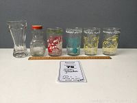 Seven vintage cartoon character glasses and jar including two Flintstones Fred hunting glasses, a Foghorn Leghorn glass, a jar with orange lid, and others.
