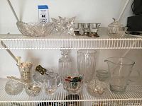 Overview of multiple crystal and glass items including cut crystal bowls, candlesticks with sterling bases, decanter, and various small glass items on white wire shelving.