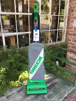Full upright green and grey Oreck XL commercial vacuum cleaner standing on an outdoor table next to plants and window.