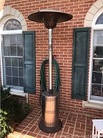Full view of the Endless Summer patio heat lamp showing tall stainless steel cylindrical base, long pole, and top heat shield against brick wall and window background.