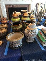 Photo showing seven ceramic vases arranged on a table, including four with lids and three with two handles. The vases feature various painted themes such as elephants and leopard spots, with some on ornate metal stands.