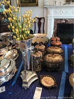 Table display showing ceramic jars with lids, silver-tone vase with faux yellow flowers, and ceramic planters with black stands.