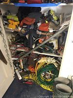 Photo showing various tools including Black and Decker power drill, extension cords, gloves, and drilling accessories piled in a small closet-like space.