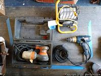Full view of the lot items on a floor mat including Ridgid sander, Ryobi drill, and yellow portable work light.