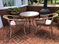 Photo showing set of round wicker table with glass top and two matching wicker armchairs with cushions in outdoor brick patio area.