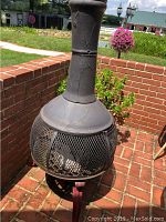 Full view of the outdoor metal fireplace with chimney in a brick patio corner, showing the cracked texture on the chimney and spherical mesh body.