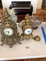 Full view of two ornate metal mantel clocks with floral ceramic bowl and other decorative items on table