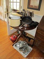 Photo showing collection of pots and pans on a small round table and on the floor, including large enamel-coated stock pot, stainless steel pots, frying pans, and glass lids in a baking pan.