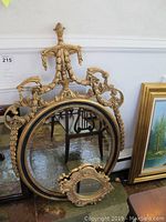 Large oval mirror with an ornate gold frame and smaller ornate oval mirror resting against it, shown with partial view of surrounding furniture.