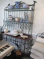 Full view of black metal bakers rack with wood top, glass shelves, and decorative blue and white ceramics on shelves to show scale and structure