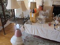 Wide view of assorted lamps, lamp shades, boxes of candles, glass cake stand with dome, and other glassware on tables