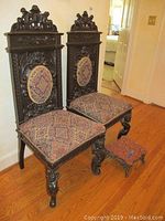 Pair of dark wood ornately carved chairs with jacquard upholstery and a small square foot stool with cast iron base on wood floor