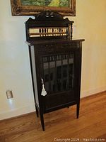Front view of wooden cabinet with glass door, carved top mirror frame, and tassel key.