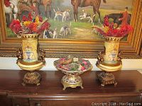 Photo showing the pair of matching ceramic vases filled with faux flowers and the decorative ceramic bowl also containing faux flowers, all placed on a wooden surface in front of a framed painting.