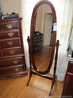 Full view of the oval floor mirror with a wooden frame and stand placed in a room with hardwood flooring near furniture.
