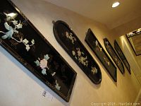 Five black lacquer Asian decorative wall panels mounted on a wall, showing a mix of rectangular and oval shapes with raised floral and bird inlays.
