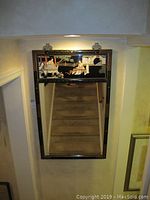 Full view of the rectangular black framed mirror hung on the wall with Asian themed painted decoration in the top panel and reflective mirror below.
