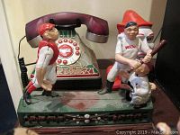 Photo showing Coca Cola corded phone with push button dialing along with baseball themed metal bank featuring batter, catcher, and umpire figures