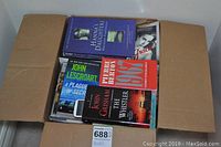 Open box showing assorted paperback books including titles by John Grisham, John Lescroart, and Laurent Binet.